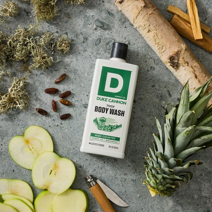 White Duke CANNON THICK BODY WASH bottle with a green ‘HIGH COUNTRY FOREST AIR + BIRCHWOOD’ label on grey stone. Surrounded by birch bark, berries, moss, green apples, a slice of pineapple and a knife.