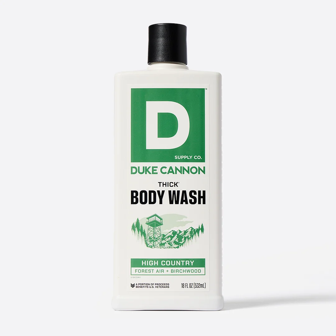 Rectangular white bottle of DUKE CANNON THICK Body Wash, 532 ml, with a black cap. Green label featuring the logo and product name. Illustration of a wooded mountain landscape with a viewing tower. Fragrance: High Country Forest Air + Birchwood.