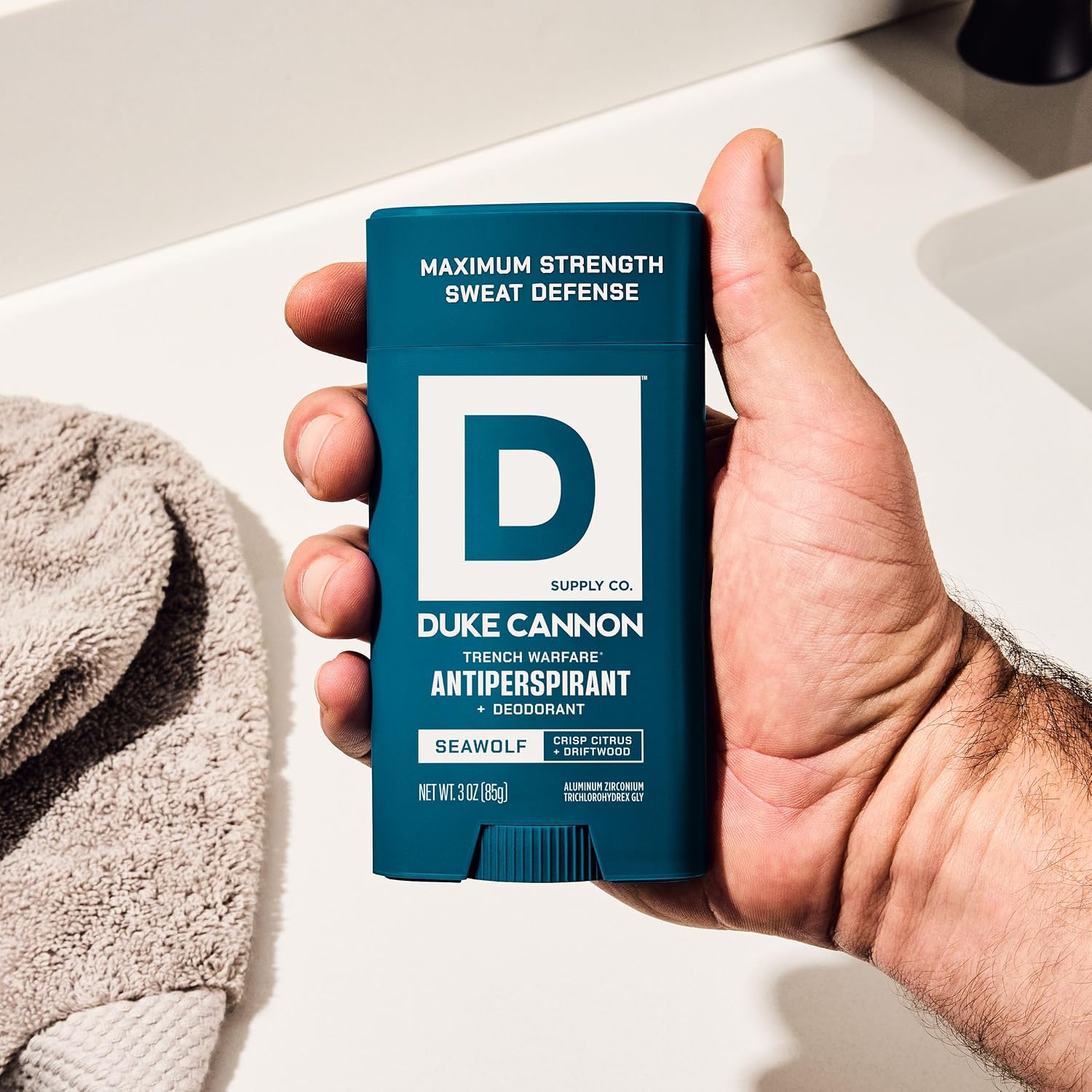 A hand holds a dark blue DUKE CANNON Trench Warfare antiperspirant deodorant stick. Front reads ‘Maximum Strength Sweat Defence’, ‘Seawolf Crisp Citrus + Driftwood’, ‘Net Wt. 3 oz (85g)’. Back: light washbasin, grey towel.