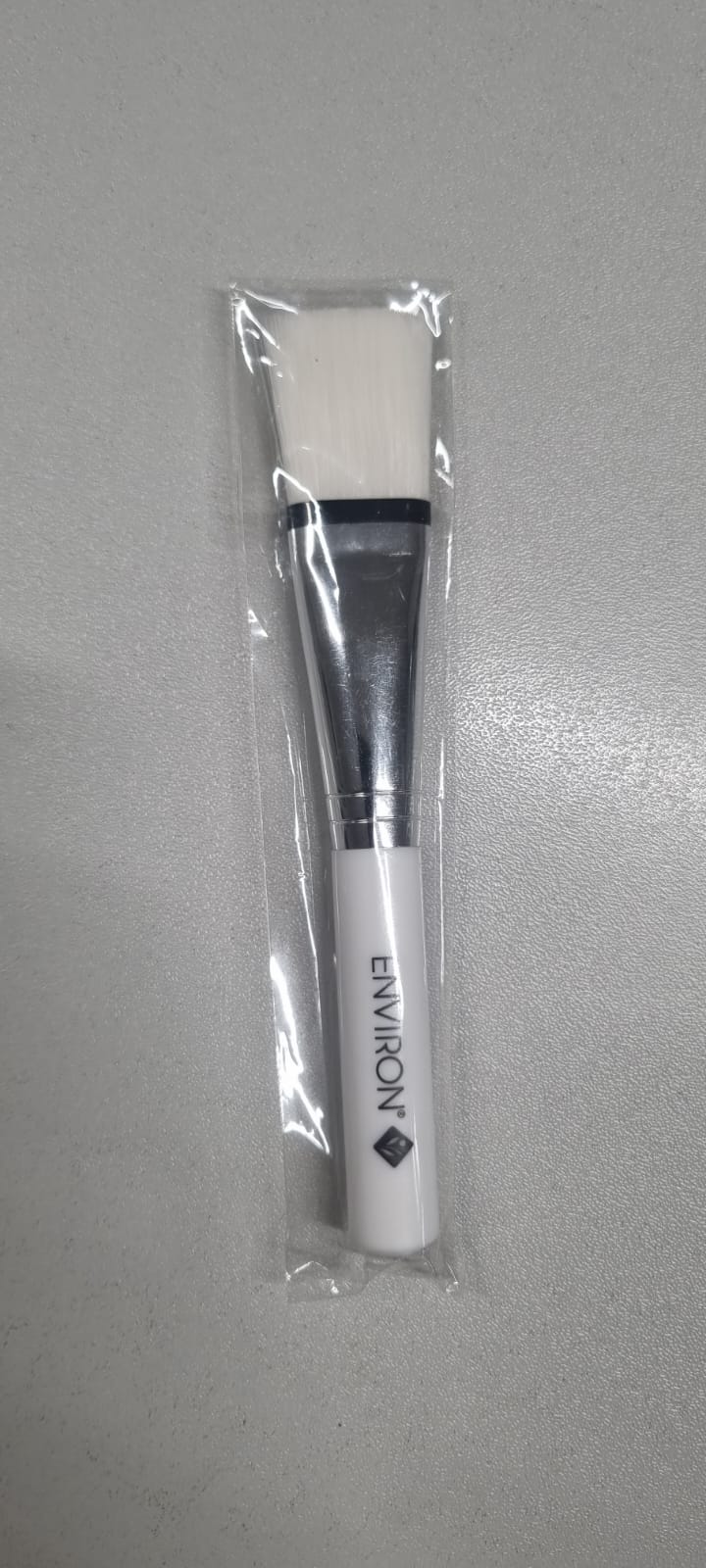 Weißer Peeling-Pinsel von Environ, in durchsichtiger Folie verpackt. Weißer Griff mit vertikalem "ENVIRON"-Logo, glänzende silberne Metallhülse und weiche, weiße, flache Borsten. Kompakt, ideal für Nagelpflege oder Reise.