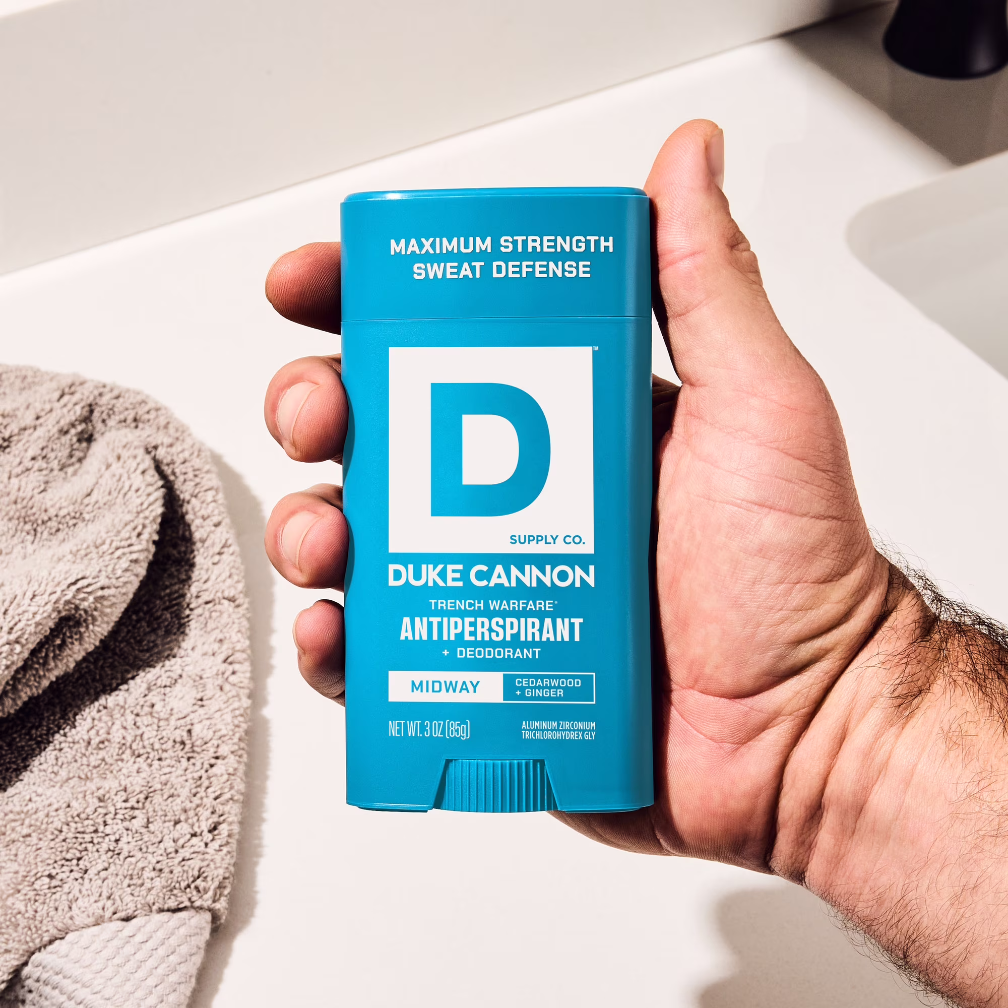 A man’s hand holds a turquoise Duke CANNON Trench Warfare antiperspirant deodorant stick. Packaging shows ‘Maximum Strength Sweat Defence’, ‘Midway Cedarwood + Ginger’ and 85g. Background: light washbasin top, grey terry towel.