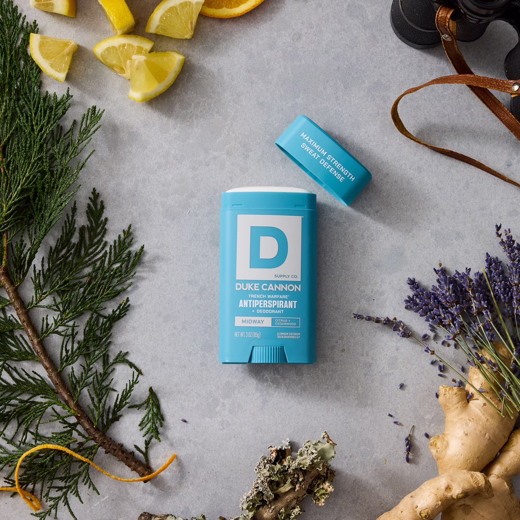 An open blue DUKE CANNON MIDWAY Trench Warfare antiperspirant deodorant stick on a grey background. Next to it, lemon slices, green cedar twigs, purple lavender flowers, ginger root, the cap and a pair of binoculars are visible.