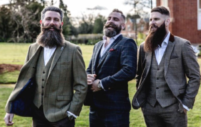 Tre uomini sorridenti con lunghe barbe curate e baffi eleganti sono all'aperto. Indossano completi in tweed a quadri con gilet e camicie bianche: a sinistra verde oliva, al centro blu scuro, a destra marrone. Sullo sfondo alberi e un edificio.