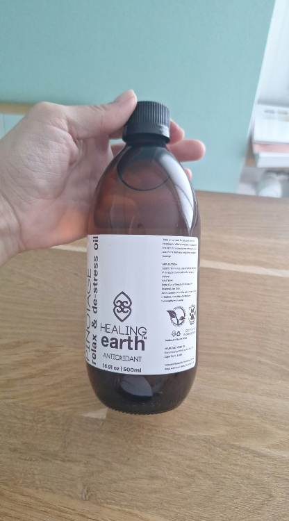 Eine Hand hält eine hohe dunkelbraune Glasflasche mit schwarzem Schraubverschluss. Auf dem weißen Etikett steht "HEALING earth", "PINOTAGE relax & de-stress oil" und "500ml". Leicht schräg über hellem Holz vor türkisblauer Wand.