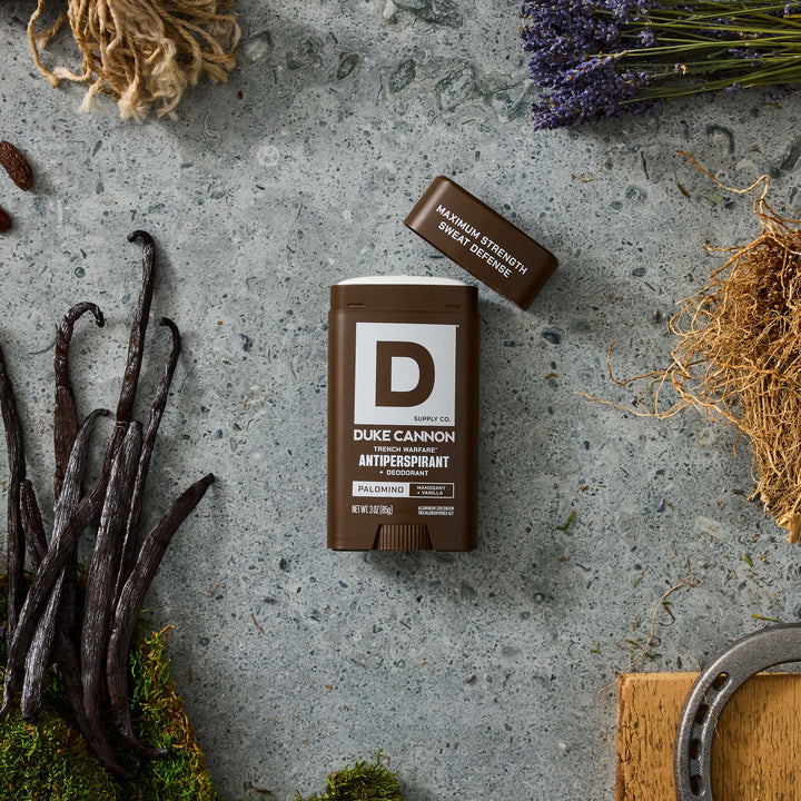A brown DUKE CANNON Palomino antiperspirant deodorant stick, without its cap, lies on a grey stone slab. It is surrounded by dried vanilla pods on green moss, purple lavender, beige ropes, straw and a horseshoe resting on a wooden block.