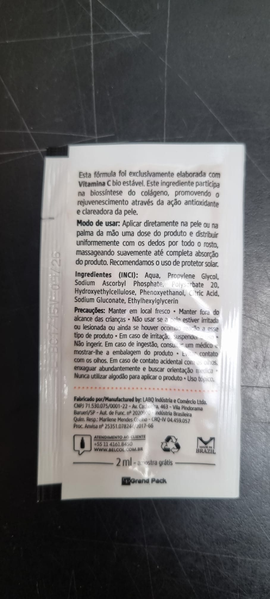 Sachet jetable blanc contenant le sérum BelCol RenoveC Fluid, texte portugais : formule stable à la vitamine C, usage visage, ingrédients, mention « échantillon gratuit 2 ml ». État : surface sombre et rayée ; légers plis.