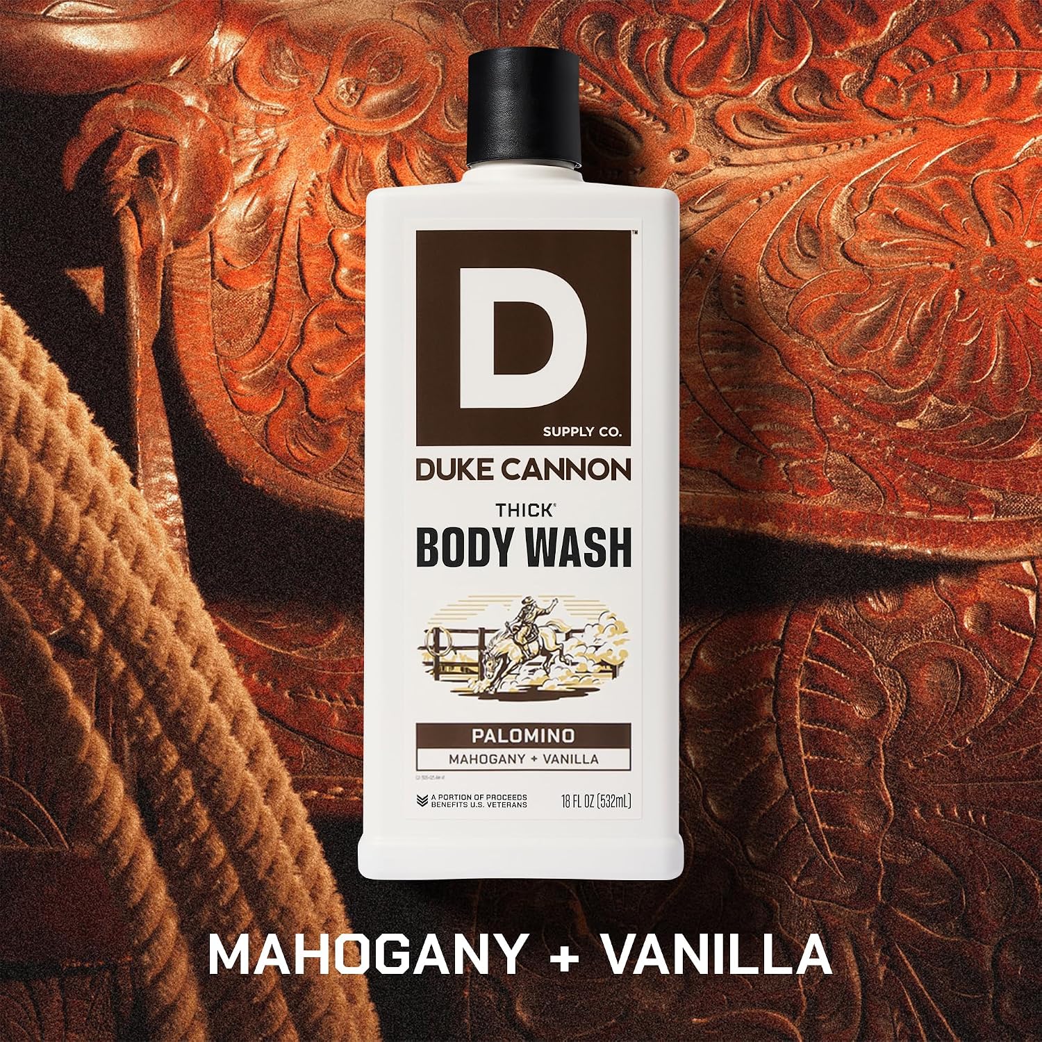 White rectangular bottle of DUKE CANNON Thick Body Wash with a black cap, Palomino Mahogany + Vanilla fragrance. Label: cowboy on a horse. The bottle stands on dark brown, richly patterned leather, resembling a saddle.