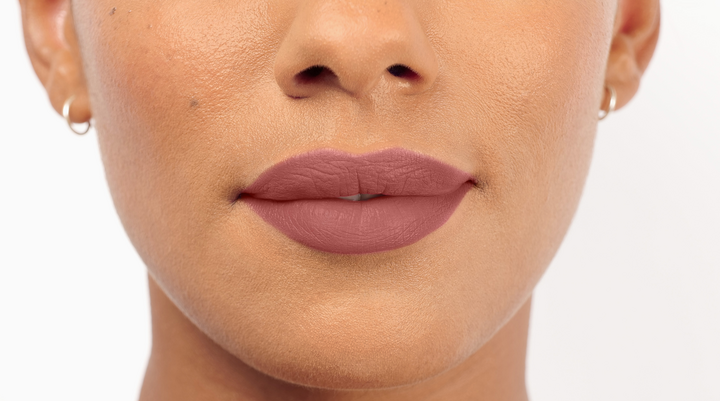 Zoom sur les lèvres et le bas du nez d’une personne au teint chaud. Les lèvres sont couvertes du rouge à lèvres Beyond Matte Lip Fixation rose poudré, au fini mat uniforme. Créoles argentées visibles.