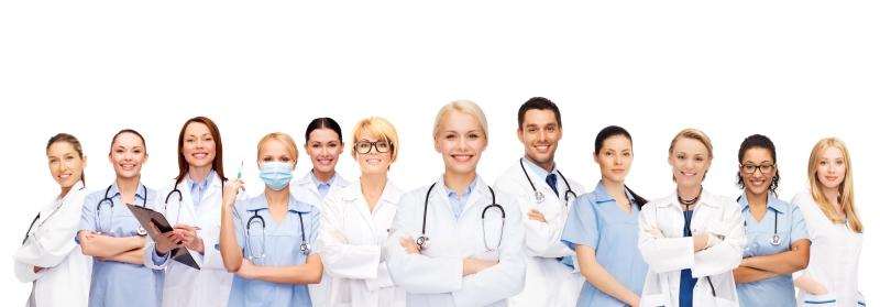 A smiling group of eleven healthcare professionals, doctors and nurses, are standing in a row. Many are wearing white coats or blue surgical scrubs with stethoscopes. Some are holding clipboards or a syringe.
