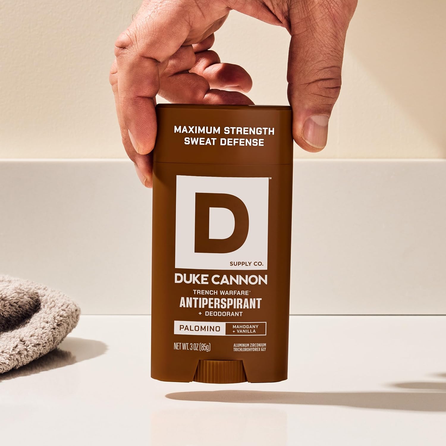 A man’s hand holds a brown DUKE CANNON Trench Warfare Palomino Antiperspirant & Deodorant against a light-coloured background. On the packaging: ‘Maximum Strength Sweat Defence’, with notes of mahogany and vanilla. 85g stick.