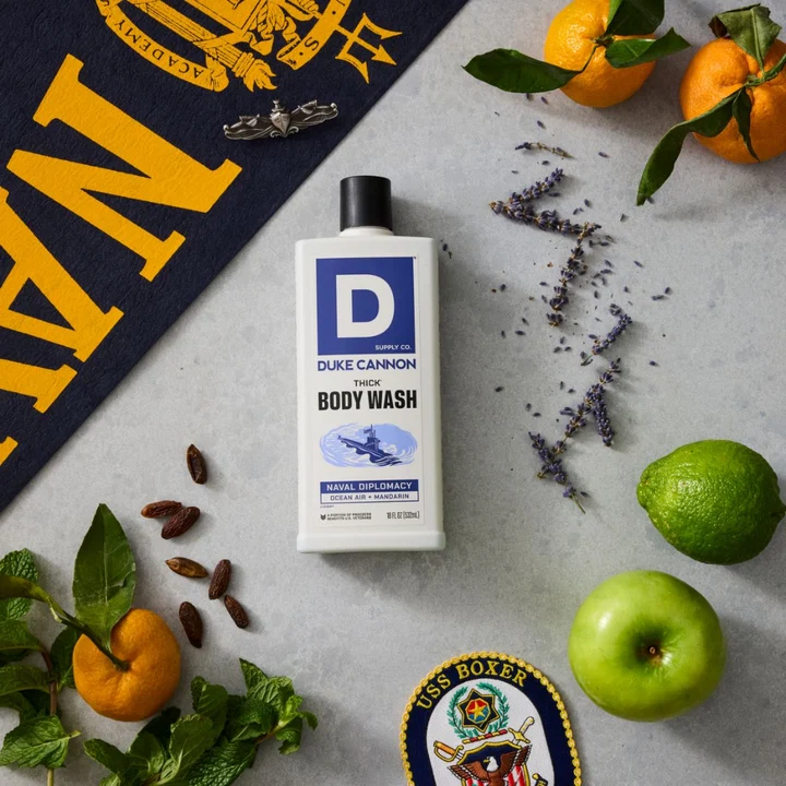 White, rectangular DUKE CANNON THICK Body Wash ‘NAVAL DIPLOMACY’ bottle featuring a blue ‘D’ logo. Enveloped by notes of mandarin, lime, green apple, mint, lavender sprigs and dates. Background: Banner and ‘USS BOXER’ patch.