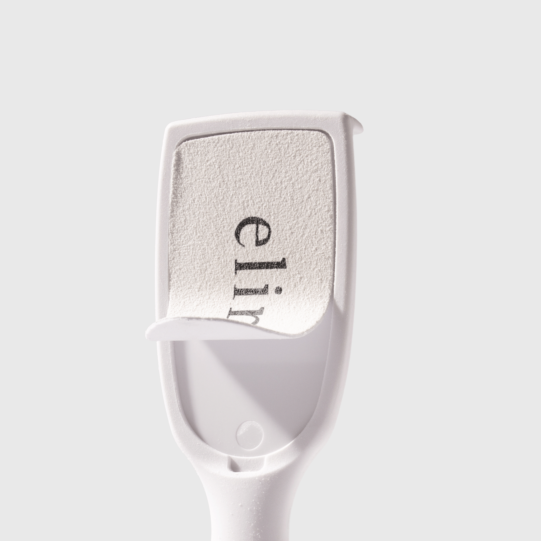 Primer plano de raspador/lima para pies ELIM Medi Ped blanco. Lado rugoso y granulado con inscripción «elim» en negro vertical. Inserto flexible ligeramente levantado; debajo, superficie lisa de plástico. Para eliminar callos.