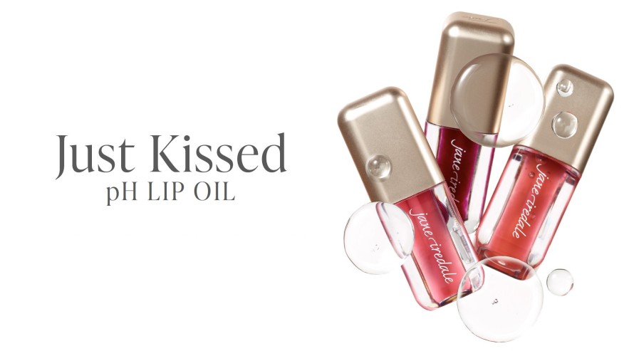 Drei Flaschen Jane Iredale Just Kissed pH Lip Oil mit goldenen Kappen und transparenten Behältern, die zartrosa bis beerig-rote Flüssigkeit zeigen. Klare, schimmernde Blasen betonen die leichte Textur. Links steht "Just Kissed pH LIP OIL".