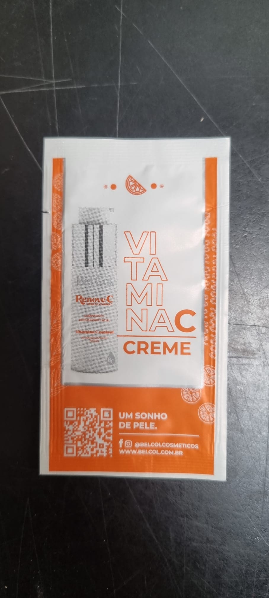 Sachet d'échantillon blanc de la crème Bel Col Renove C, motif orange aux bords et oranges stylisées. En haut, symbole vitamine C, puis « VITAMINA C CREME ». En bas, zone orange : QR code, « UM SONHO DE PELE. » et réseaux sociaux.