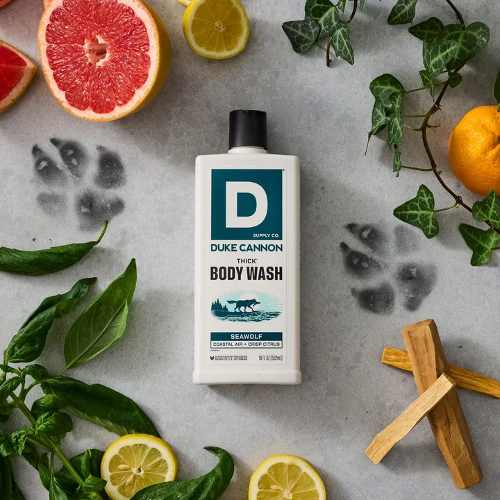 A white bottle of DUKE CANNON SEAWOLF THICK Body Wash on a grey background. Surrounded by citrus fruits, green leaves and light-coloured pieces of wood. Two faint paw prints emphasise fresh, aquatic and woody notes.
