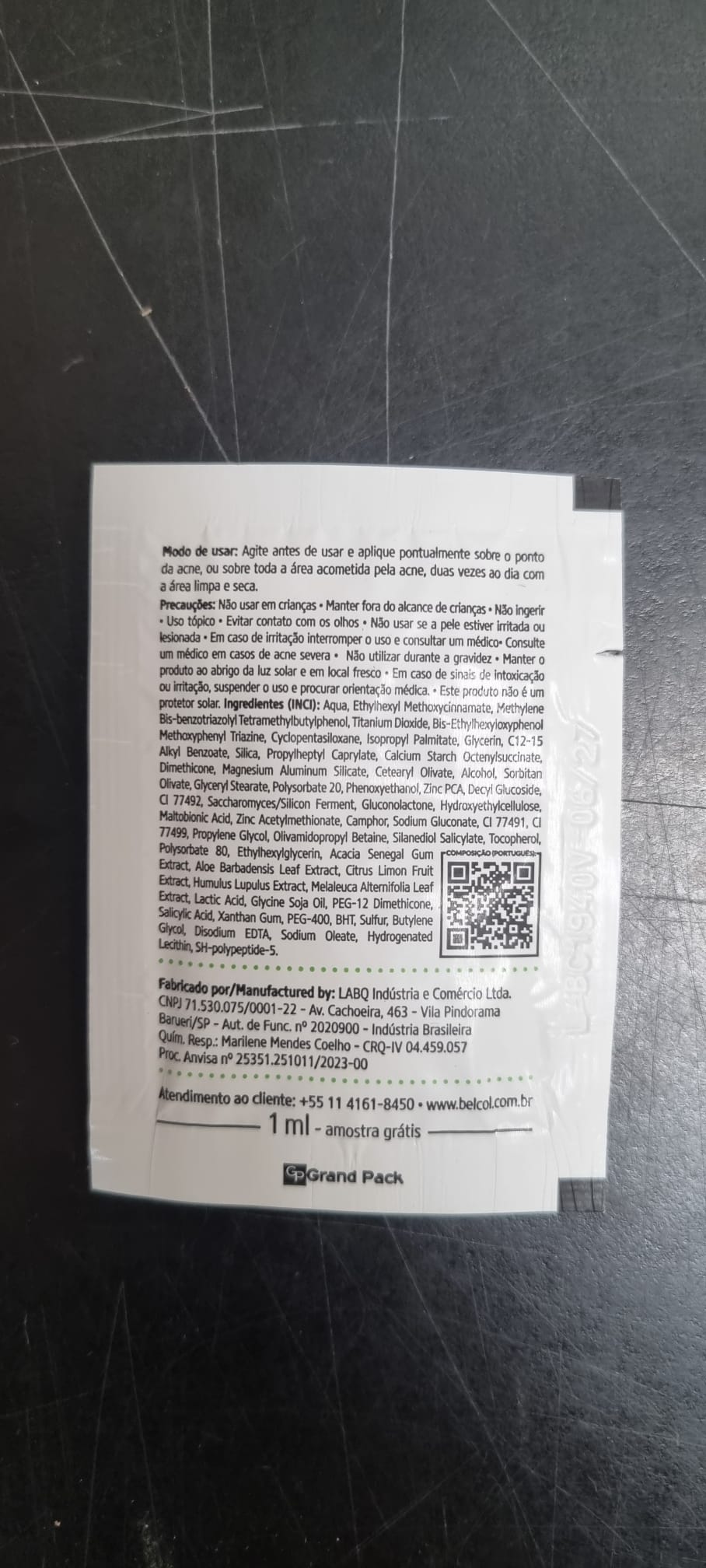 Sachet blanc d'échantillon 1 ml de lotion de jour anti-acné Tri-Protect BelCol, avec protection UV SPF 45. Mode d'emploi et ingrédients (zinc PCA, soufre), infos fabricant en portugais. QR code visible. Surface sombre, légèrement rayée.