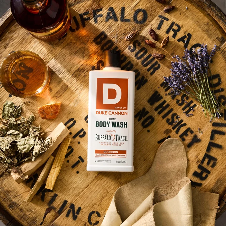 Flacon blanc de gel douche Duke CANNON Bourbon Thick Body Wash sur un tonneau. À côté : verre de bourbon, bouteille de whisky, lavande, bois de cèdre et ambre, évoquant ses notes parfumées.