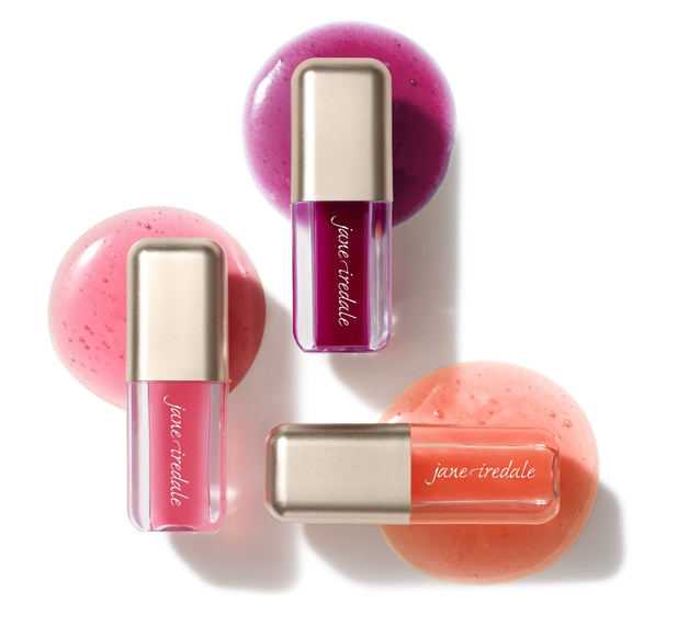 Drei längliche Lipgloss-Tuben von Jane Iredale liegen auf weißem Untergrund. Sie enthalten glänzendes Produkt in Tiefmagenta, Pink und Korallorange und liegen jeweils auf einem Klecks ihres Farbtons. Goldene Kappen, Logo sichtbar.
