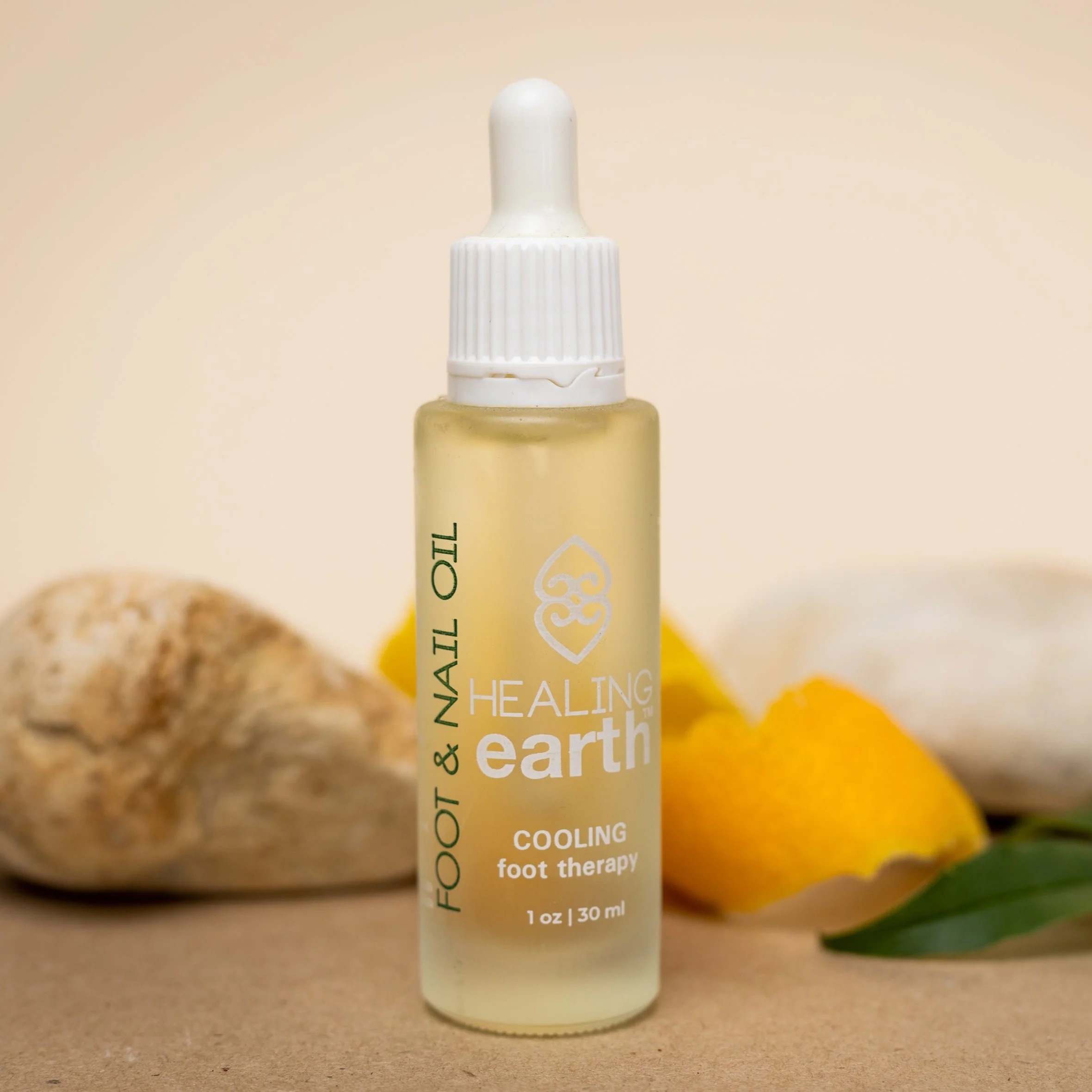 Transparente Pipettenflasche mit gelblichem Cooling Foot & Nail Oil von Healing Earth, beschriftet mit „FOOT & NAIL OIL“ und „30 ml“. Auf beigem Untergrund, umgeben von hellen Steinen, Zitrusschale und grünem Blatt. Szene wirkt natürlich und pflegend.