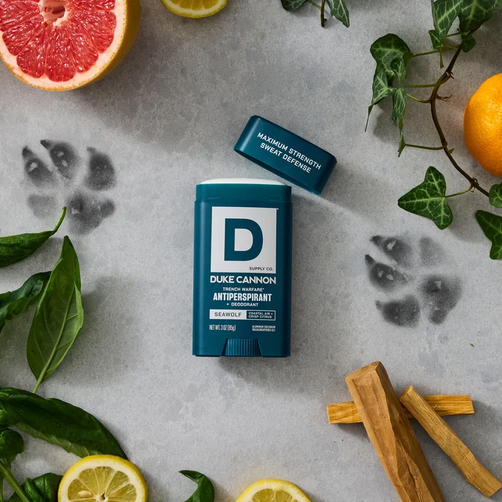 A dark blue Duke CANNON Seawolf antiperspirant deodorant with its lid open, set against a light grey background. Scattered around it are slices of grapefruit and lemon, green leaves, pieces of wood and dark paw prints.