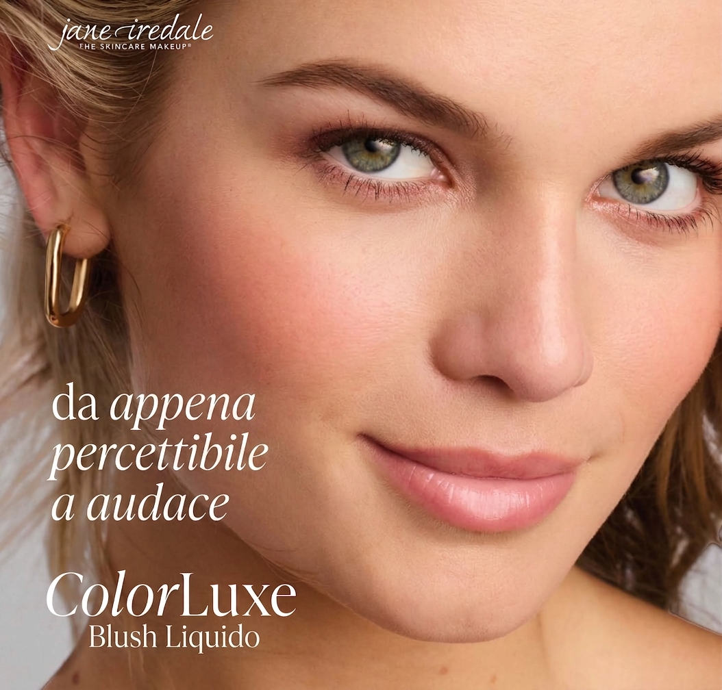 Primo piano di una donna con la pelle leggermente luminosa e un delicato fard rosa, che guarda verso la fotocamera. Occhi verdi delicatamente sottolineati, orecchino dorato a forma di U. Messa a fuoco sul centro del viso, look trucco fresco e naturale.